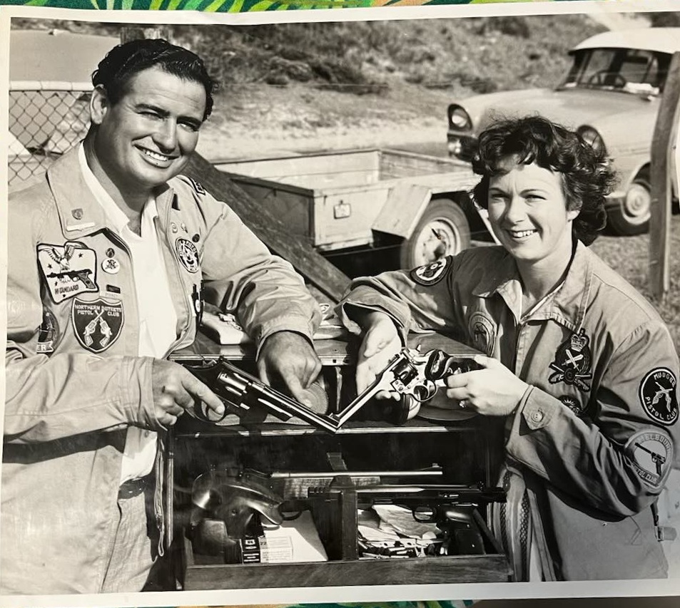 A man and a woman in patch-covered shooting jackets comparing revolvers over an open gun case at an outdoor range