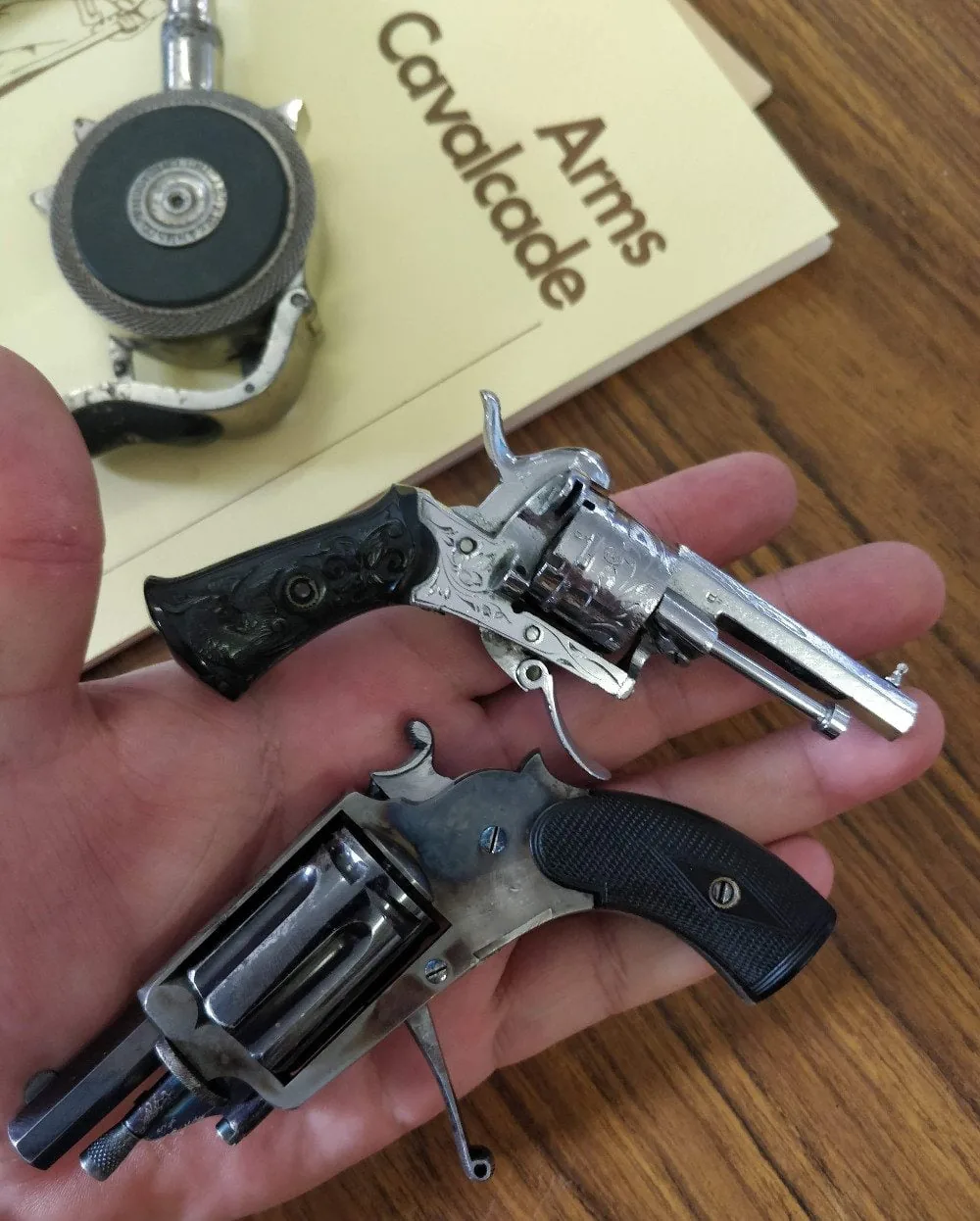 Two nineteenth-century pinfire revolvers held in one hand, beside a book titled Arms Cavalcade
